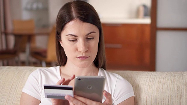年轻女子坐在客厅的沙发上用智能手机刷卡在线购物视频素材
