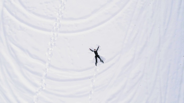 从空中俯瞰，一个男人穿着雪鞋躺在白雪覆盖的田野上做雪天使。人们喜欢并享受户外的乐趣。4k无人机飞行直线向下的视角视频素材