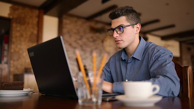 英俊的自由职业者，戴着眼镜，在咖啡馆用笔记本电脑努力工作。一个人在键盘上打字，在网吧上网找新工作。经营理念视频素材