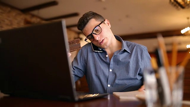 哈里英俊的年轻商人戴着眼镜在笔记本电脑上工作，打电话，做笔记，在笔记本上搜索信息，喝咖啡和吃蛋糕。多任务telecomuting概念视频下载