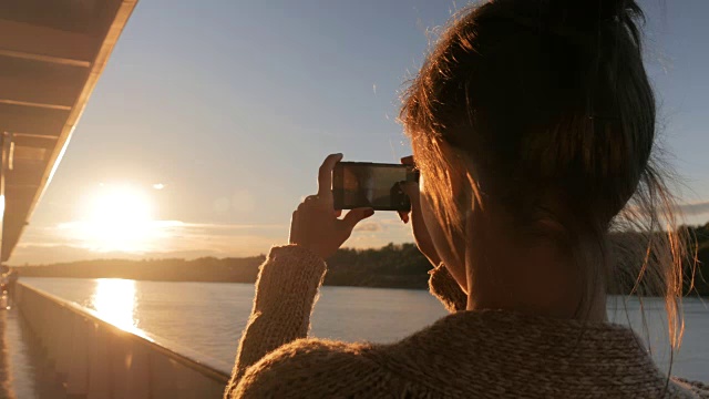 一名女子在游轮甲板上用智能手机拍摄日落剪影视频素材
