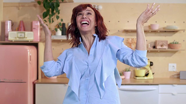 一位红头发的中年妇女站在厨房里，双手穿过头发，微笑，大笑，高举双手。视频下载