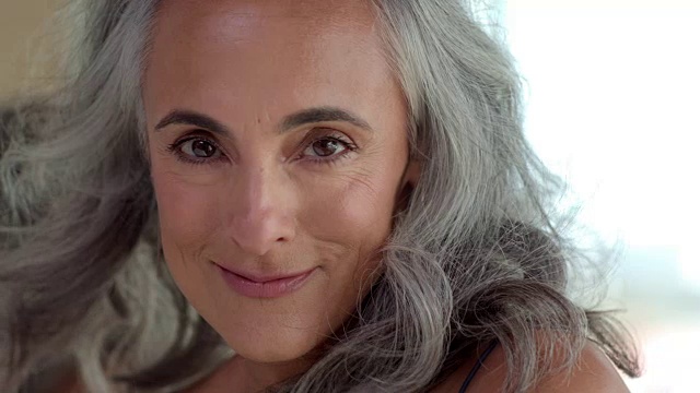 一个中年妇女的特写，灰色的头发看着和微笑的相机，头发从风吹来，对模糊的背景。视频素材