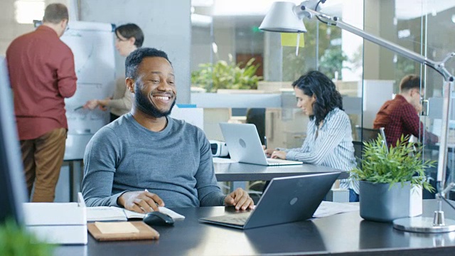 一名黑人在办公桌前用笔记本电脑工作时，遇到了有趣的错误并大笑。有创造力的年轻人在做他们的工作。现代明亮的办公室。视频素材