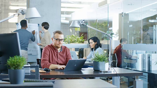 忙碌的国际公司办公室，白种人在他的办公桌上用笔记本电脑工作。在背景创造性的年轻人工作。视频素材