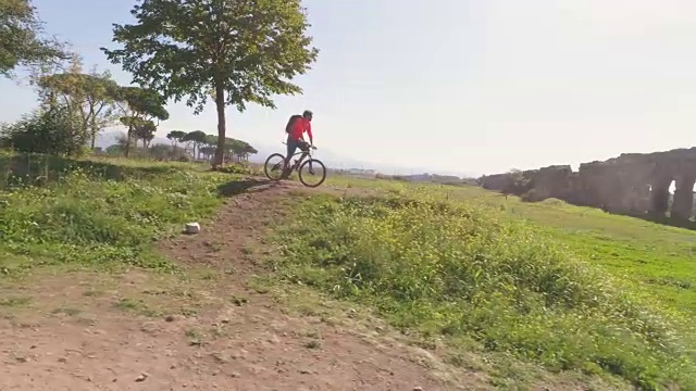 年轻的骑自行车的人骑在土路到达一个小山和停在树前的古罗马渡槽在橙色运动服和背包鸟瞰无人机手推车视频下载