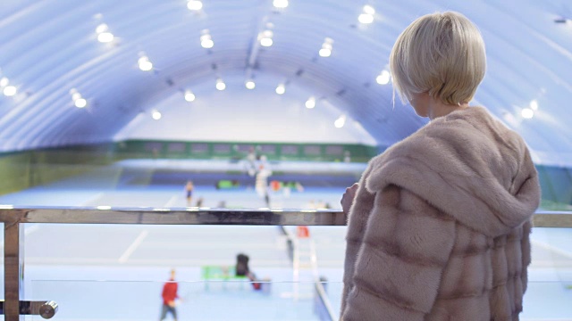 穿着外套的年轻女子在球场的阳台上观察网球运动员视频下载