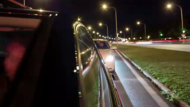 一辆开着的德国黑色轿车在夜间长时间曝光延时拍摄，相机背对着行驶方向，前景中窗户有反光，背景交通是运动模糊，男性司机部分透过挡风玻璃看到视频素材