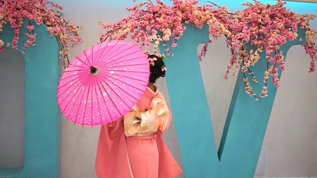 穿着粉红色和服的艺妓带着雨伞在墙边走着，上面写着樱花的大字母。视频下载