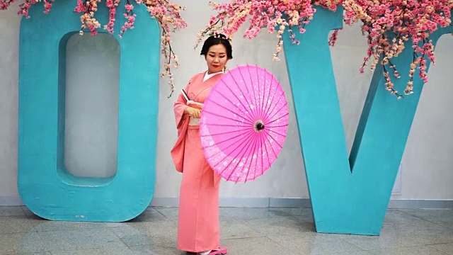 艺伎穿着粉红色的衣服在墙边走着，大爱带着樱花和转动着雨伞。视频下载