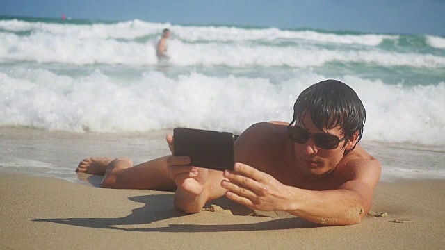 男游客拿着智能手机在海边的海滩上自拍。慢动作浪花飞溅。1920x1080视频下载