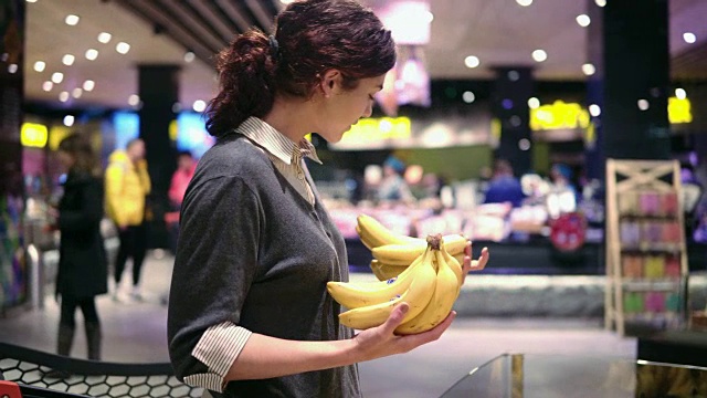 年轻漂亮的黑发女孩在二十多岁的时候，在一家杂货店的水果和蔬菜通道里挑香蕉，把它们放进购物车视频素材