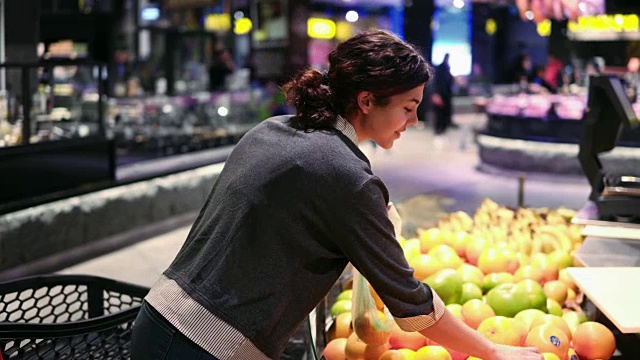 年轻漂亮的黑发女孩，20多岁，在杂货店的水果蔬菜货架上，从塑料袋里取出橙子。视频素材