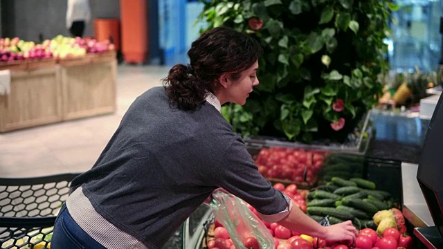 年轻漂亮的黑发女孩，20多岁，在杂货店的水果蔬菜货架上挑着装在塑料袋里的西红柿视频素材
