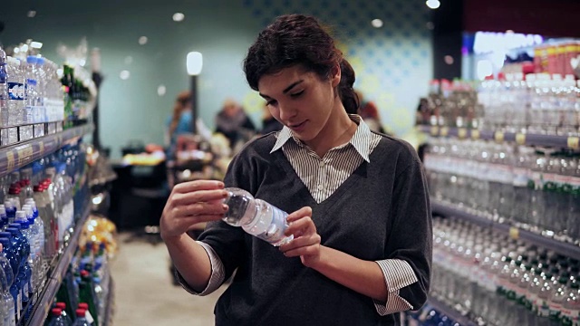 年轻漂亮的黑发女孩在她的20多岁仔细看水瓶标签，试图决定买哪一个。在杂货店购物视频素材