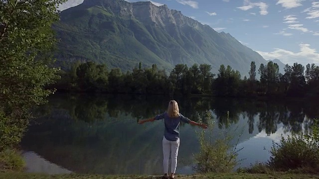 无人机拍摄到一名年轻女子站在湖边双臂张开，无人机飞了过来视频下载