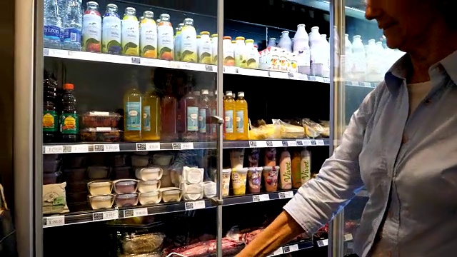 一位面目人非的老妇人在食品市场购物，从冰箱里拿出一件商品，放进购物车里视频下载