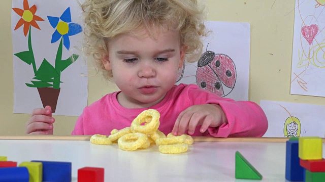 可爱的脸女孩吃玉米圈坐在她的桌子旁边玩具和画视频下载