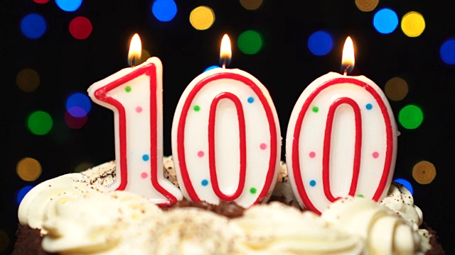 蛋糕上的100号——100支生日蜡烛燃烧着——最后吹灭。颜色模糊的背景视频下载