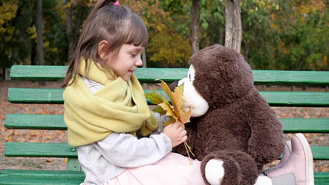 在公园的操场上，孩子们坐在长椅上，手里拿着黄叶，玩着泰迪熊玩具视频素材