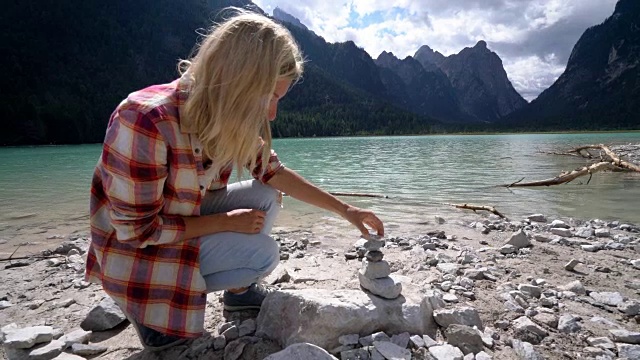 年轻女子在湖边堆石头。女人站在令人惊叹的高山湖泊附近在白云石阿尔卑斯山脉制造一堆石头与她的手视频素材