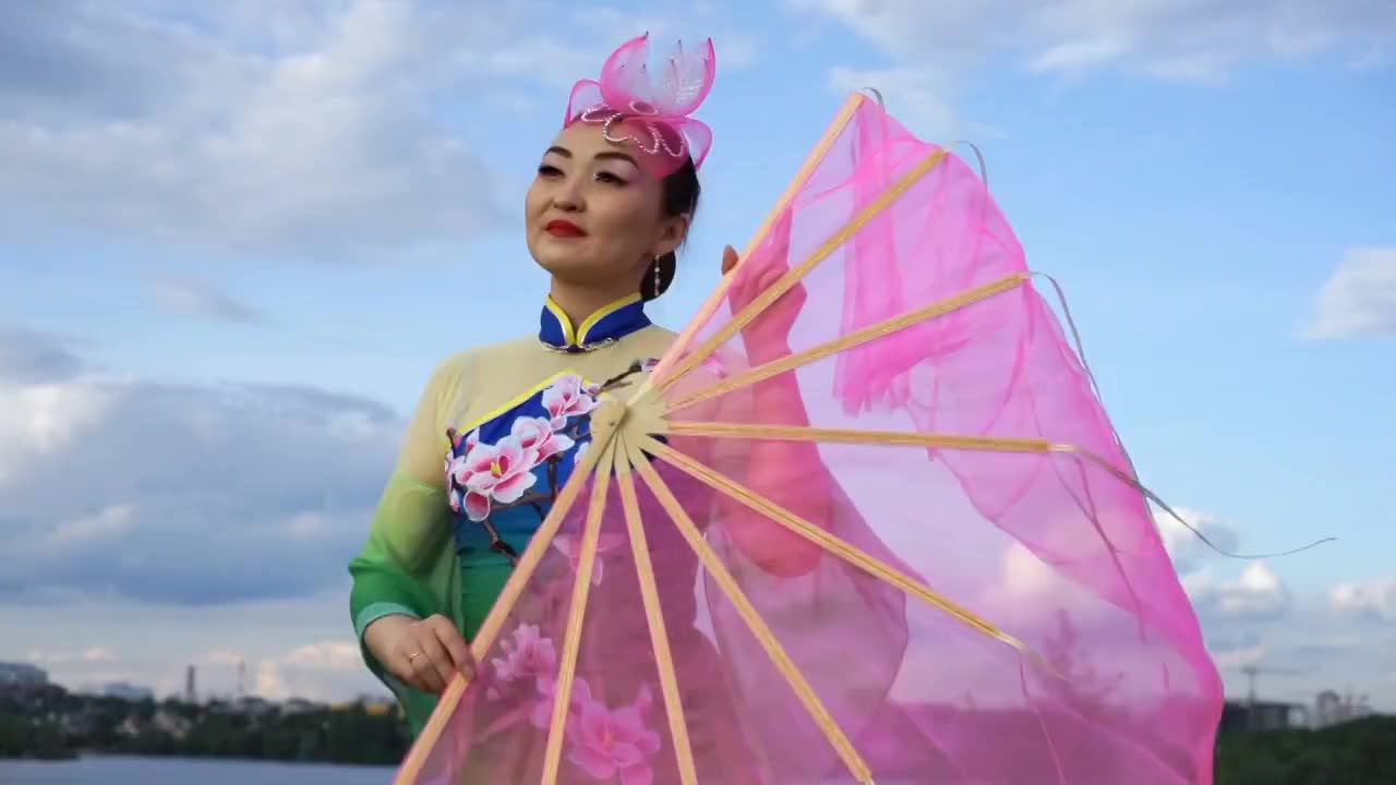 美丽的中国女孩穿着中国传统服装，拿着大粉扇在河岸上跳舞视频素材