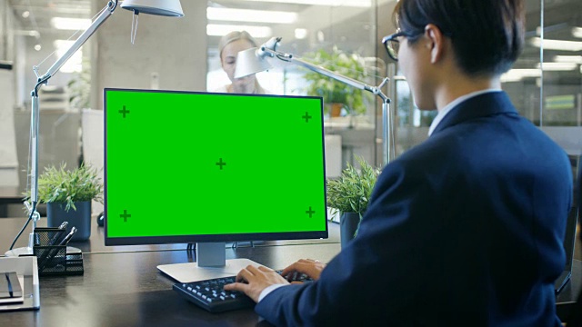 在办公室里，商人在他的办公桌上用一台模拟的绿色屏幕的个人电脑工作。同事走进办公室，并在她的办公桌前发生。视频素材