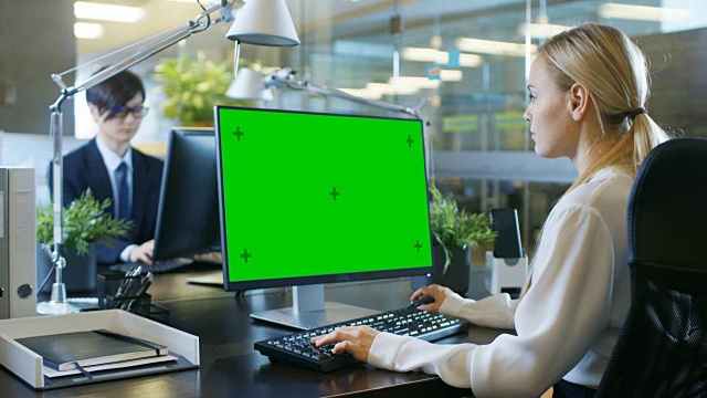在办公室里，女商人在她的办公桌前对着一台模拟的绿色屏幕的个人电脑工作。同事走进办公室，并在他的办公桌前发生。视频素材