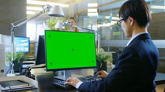 在办公室里，商人在一台模拟绿色屏幕的台式个人电脑上工作。同事走进办公室，并在她的办公桌前发生。视频素材