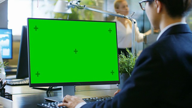 在办公室里，商人在一台模拟绿色屏幕的台式个人电脑上工作。同事走进办公室，并在她的办公桌前发生。视频素材
