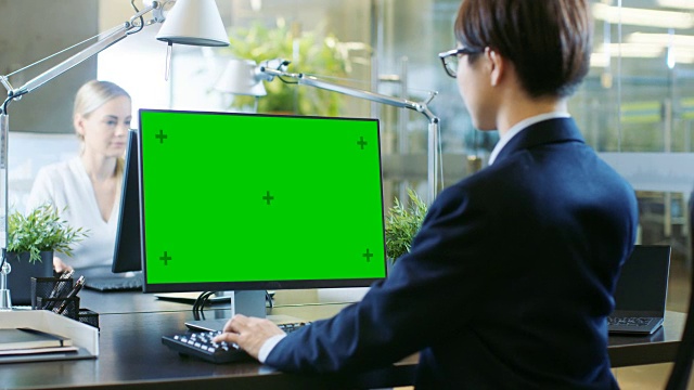 在办公室里，东亚商人在一台模拟绿色屏幕的台式个人电脑上工作。他戏剧性地转头，越过肩膀看向镜头。同事走进办公室，并在她的办公桌前发生。视频素材