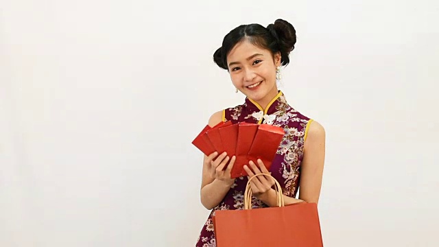 年轻的亚洲美女穿着旗袍，在她的购物袋里发现了红包为中国新年活动的白色背景。假日和生活方式的概念。旗袍衣服视频下载