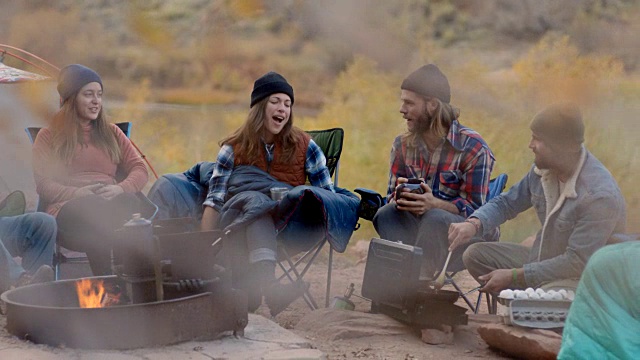 一群露营的朋友在篝火边谈笑风生，年轻人正在煎锅里炒鸡蛋。视频素材