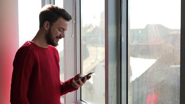 年轻快乐的男人在窗口用手机发短信。视频素材