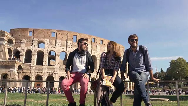 三个年轻的朋友观光客坐在罗马斗牛场前谈论在君士坦丁拱门附近玩的背包太阳镜快乐美丽的女孩长头发视频素材