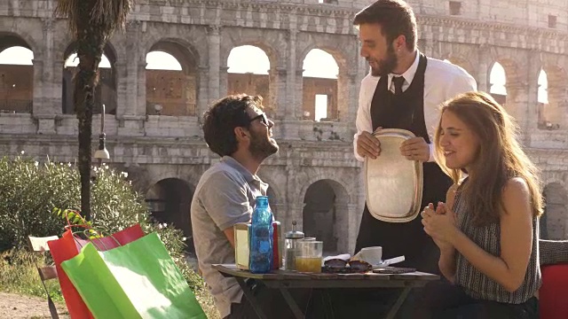 快乐的年轻夫妇游客聊天风趣的意大利优雅的服务员坐在酒吧餐厅前罗马斗牛场在日落开玩笑和大笑，有乐趣视频素材