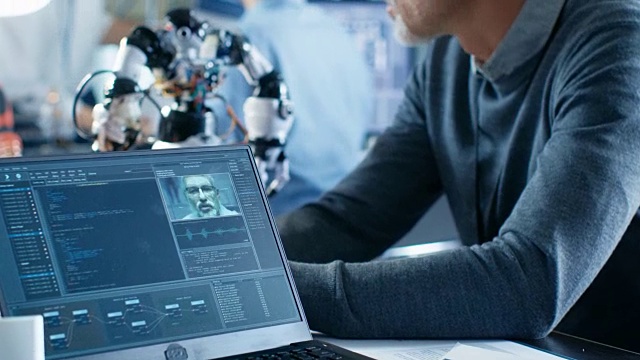 机器人工程师操纵语音控制机器人，笔记本电脑屏幕显示语音和人脸识别软件。背景机器人研究中心实验室。视频素材