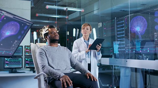 在实验室里，戴着脑波扫描耳机的人坐在椅子上，而科学家用平板电脑监督过程。在现代大脑研究/神经研究实验室。监视器显示脑电图阅读和图形大脑模型。视频素材