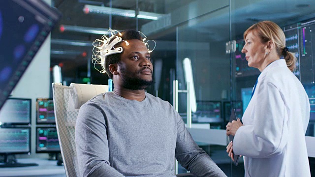 戴着脑电波扫描耳机的人坐在椅子上，而科学家用平板电脑监督过程。在现代大脑研究实验室监测显示脑电图阅读和大脑模型。视频素材