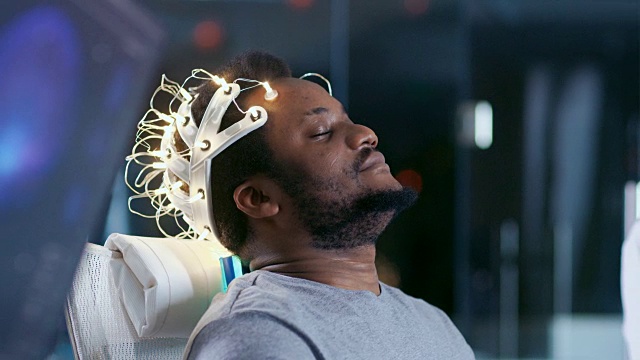 在实验室里，戴着脑电波扫描耳机的人闭着眼睛坐在椅子上。监视器显示脑电图阅读和图形大脑模型。在现代大脑研究/神经研究实验室。视频素材