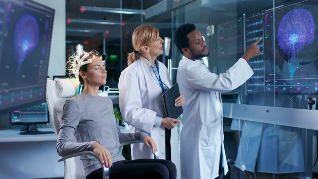 戴着脑电波扫描耳机的女人坐在椅子上，两名科学家在监督和查看数据。在现代大脑研究实验室监测显示脑电图阅读和大脑模型。视频素材