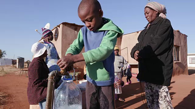 在南非，由于严重的干旱，一个非洲小男孩从水龙头里取水，而一个妇女在排队用塑料容器取水视频素材