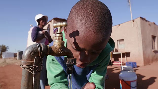 在南非，由于严重的干旱，年轻的非洲男孩从水龙头喝水，而妇女排队收集塑料容器中的水视频素材