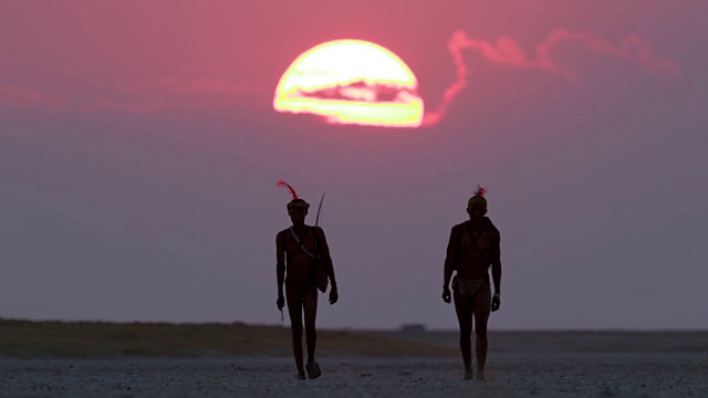 博茨瓦纳，两名圣布须曼人正穿过Makgadikgadi pan，身后是夕阳西下的红色天空视频素材