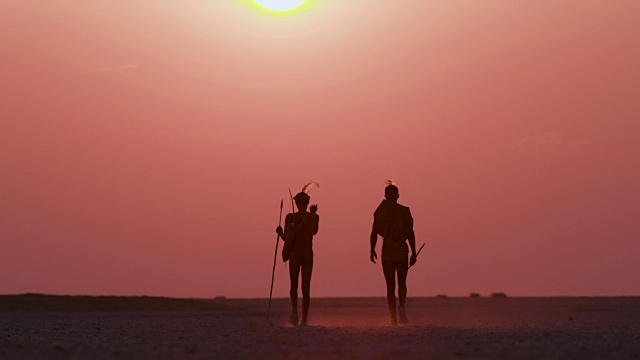 在博茨瓦纳的Makgadikgadi pan上，两名圣布须曼人走向夕阳下的红色天空的后视图视频素材