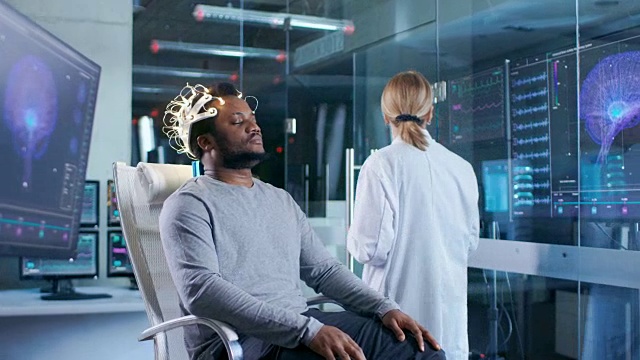 戴着脑电波扫描耳机的人坐在椅子上，而科学家用平板电脑监督过程。在现代大脑研究实验室监测显示脑电图阅读和大脑模型。视频素材