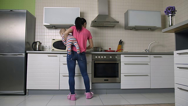 忙碌的妈妈抱着婴儿在厨房做饭视频素材