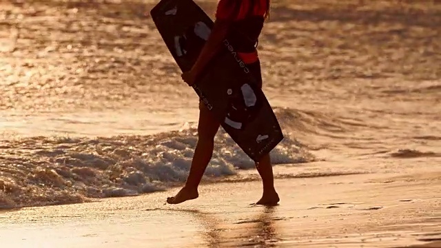SLO MO女人在海滩上拿着她的空中风筝和板在另一只手，太阳正在下山视频下载