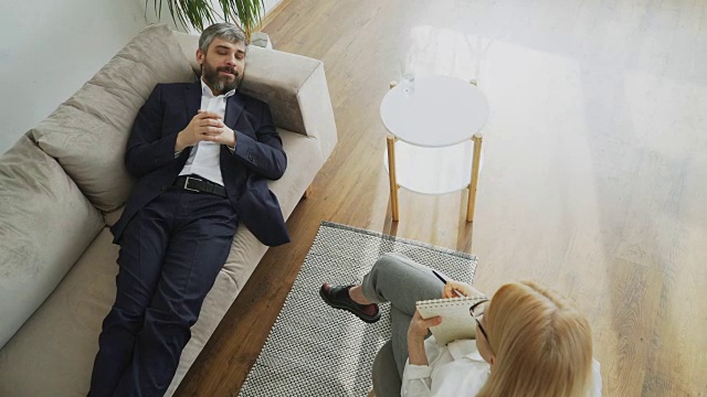 俯视男人躺在沙发上谈论他的问题与专业的女性心理治疗师或心理学家在她的办公室视频素材
