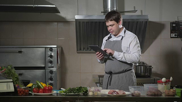 年轻的厨师看着平板电脑。在餐馆的厨房里做饭视频下载